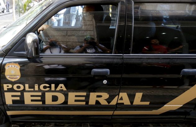 Carro da Polícia Federal com reflexos de pessoas e fotógrafos nos vidros, incluindo um com bandeira do Brasil.