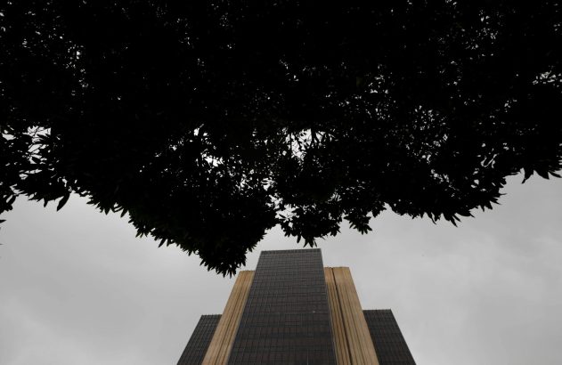 Um edifício alto visto de baixo, emoldurado por folhagem escura contra um céu nublado.