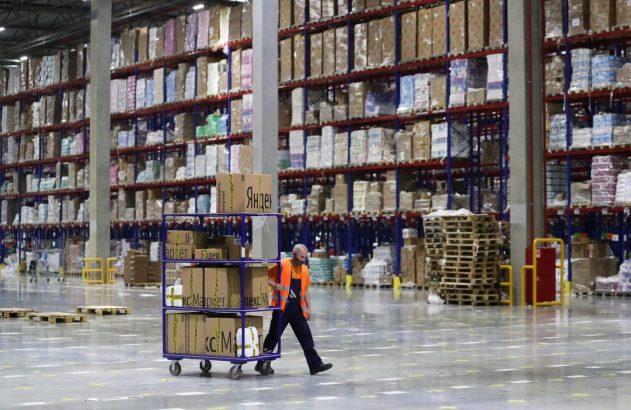 Funcionário empurra carrinho com caixas da Yandex Market em um amplo armazém com prateleiras cheias de produtos.