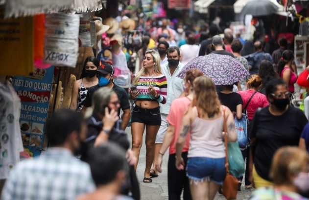 Multidão em rua comercial. Mulher com camiseta colorida, homem de máscara e outras pessoas.