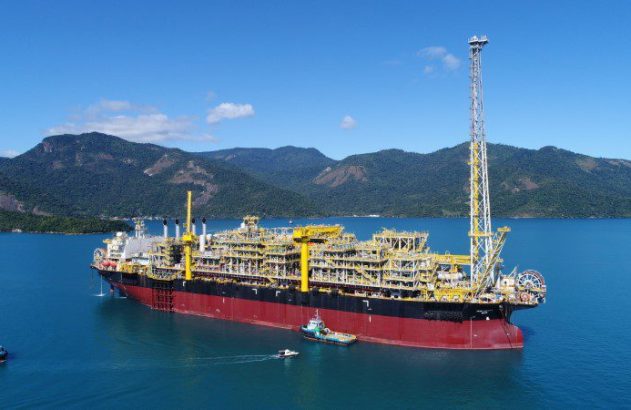 Navio FPSO vermelho e preto com vastas estruturas industriais amarelas em mar azul. Montanhas verdes e rebocadores próximos.