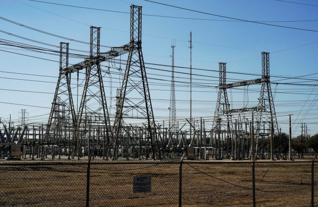 Subestação elétrica com inúmeras torres de transmissão, linhas de alta tensão e equipamentos. Há antenas ao fundo e cerca em primeiro plano.