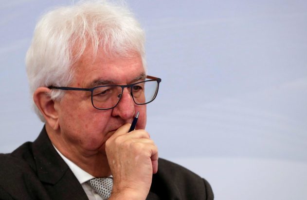 Homem de óculos, cabelo branco, em terno e gravata, segura caneta no rosto, com expressão pensativa.