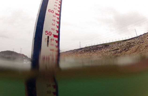 Medidor de nível de água com marcação 48, parcialmente submerso. Ao fundo, paisagem rochosa e cabos de energia.