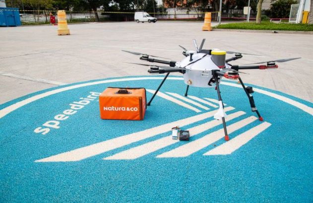 Drone de entrega multirotor pousado em plataforma azul com logo Speedbird, ao lado de pacote Natura & Co.
