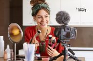 Mulher sorri e mostra pincéis de maquiagem para câmera, gravando vídeo de beleza em casa, com produtos na mesa.