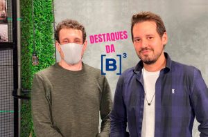 Dois homens posam para 'DESTAQUES DA [B]³'. Um usa máscara, o outro tem barba, em frente a uma parede verde.