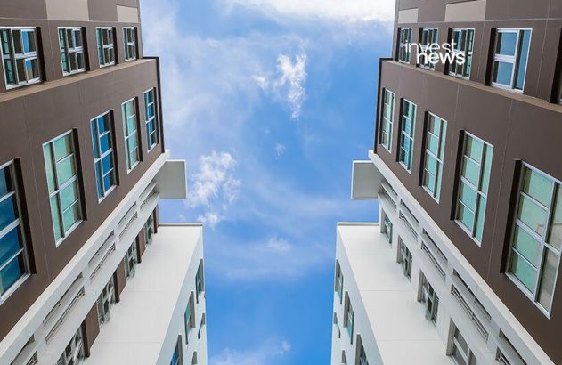 Dois prédios altos vistos de baixo, emoldurando o céu azul com nuvens brancas.