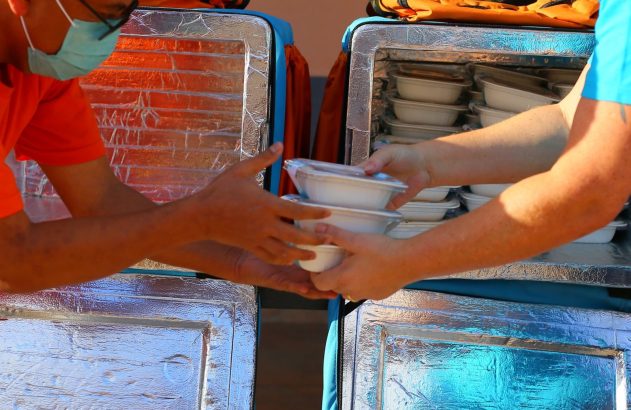 Uma pessoa entrega potes de comida a outra, ambas de máscara, usando grandes caixas térmicas.