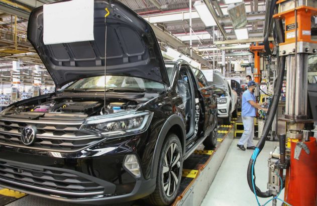 Carro VW preto com capô aberto em linha de montagem industrial. Trabalhador opera maquinário ao lado.