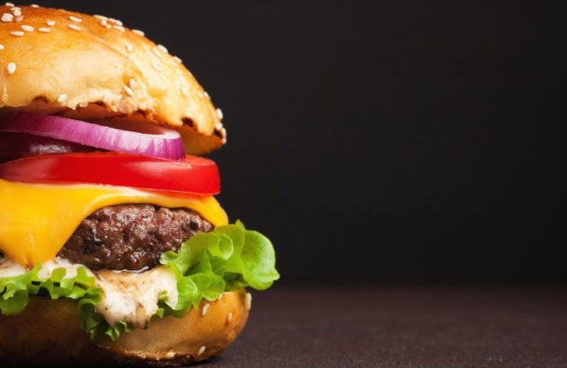 Hambúrguer com pão de gergelim, carne, queijo derretido, tomate, cebola roxa, alface e molho, em fundo escuro.