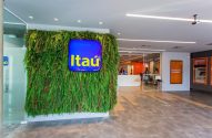 Interior de agência Itaú com parede verde, logo, recepção, caixas eletrônicos e pessoas circulando.