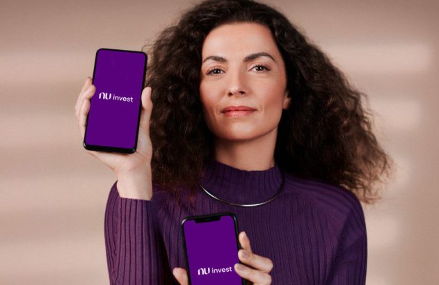 Mulher segura dois smartphones com logo 'nu invest' em tela roxa. Ela usa suéter roxo e olha para a câmera.