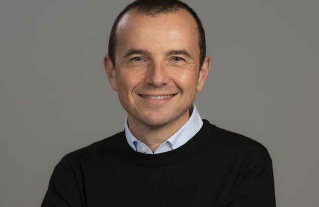 Retrato de meio-corpo de um homem sorrindo, com suéter preto e camisa azul listrada, em fundo cinza.