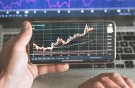 Mãos seguram smartphone exibindo gráfico de mercado financeiro, com tela de laptop mostrando gráficos maiores ao fundo.