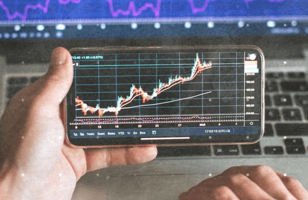 Mãos seguram smartphone exibindo gráfico de mercado financeiro, com tela de laptop mostrando gráficos maiores ao fundo.