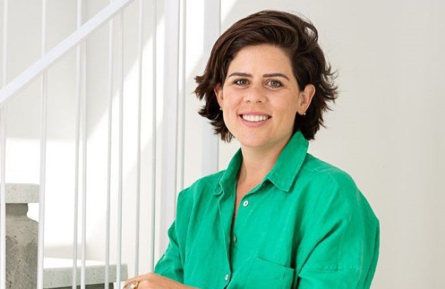 Mulher sorrindo, cabelo escuro e camisa verde. Fundo branco com corrimão.
