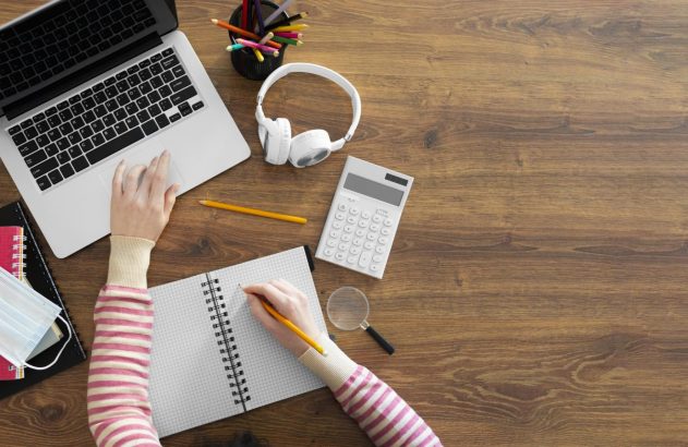 Pessoa trabalhando com laptop, escrevendo em caderno, em mesa com calculadora, fones e máscara.
