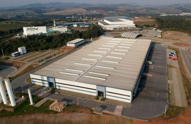 Vista aérea de um complexo industrial com grande armazém, pátio de carga e outras fábricas ao fundo.