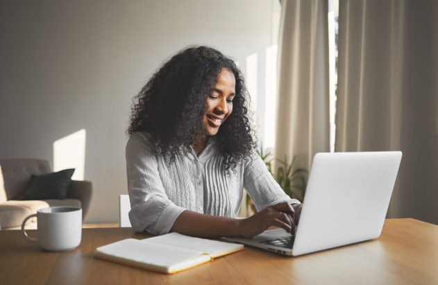 Mulher jovem sorrindo, digitando em laptop em mesa de madeira com caderno e caneca.