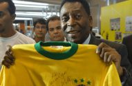 Pelé sorrindo, segurando uma camisa amarela da seleção brasileira autografada.