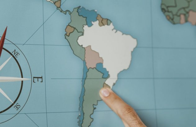 Mão apontando para um país na América do Sul em um mapa. Uma rosa dos ventos com 'NE', 'E', 'SE' é visível.