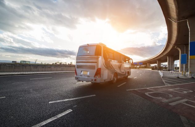 Ônibus prateado em uma estrada, passando sob um viaduto curvado, com sol forte ao fundo.
