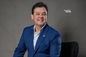 Homem sorrindo, de terno azul e camisa xadrez, sentado. Fundo cinza.