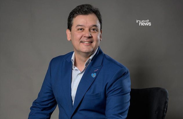 Homem sorrindo, de terno azul e camisa xadrez, sentado. Fundo cinza.