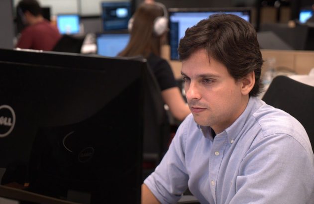 Homem focado no computador em escritório com colegas ao fundo.