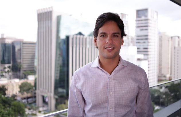 Homem sorrindo de camisa quadriculada, em varanda com prédios da cidade ao fundo.