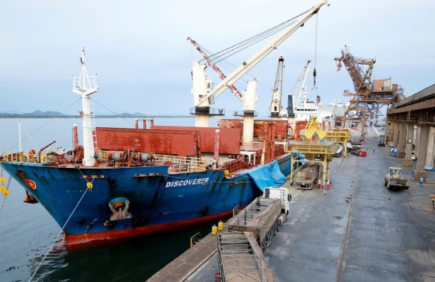 Navio cargueiro 'Discoverer' em porto, sendo carregado por guindastes e transportador para caminhões.