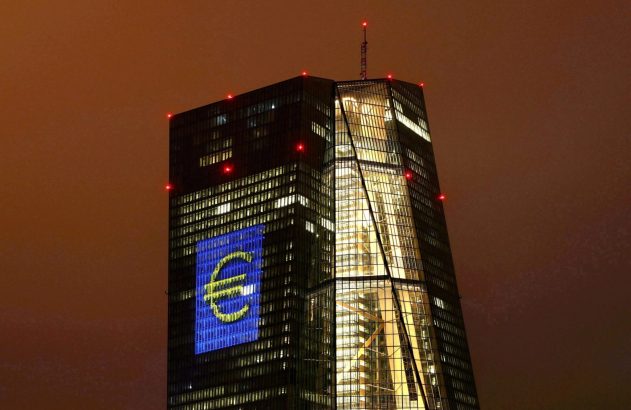 Edifício moderno iluminado à noite, com o símbolo do Euro em azul e amarelo projetado em sua fachada.