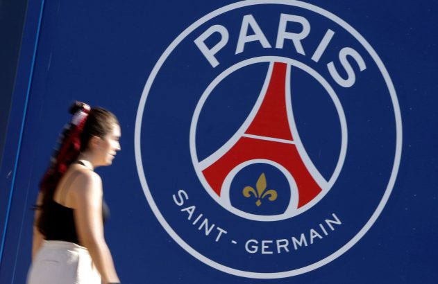 Mulher desfocada passa por grande logotipo do Paris Saint-Germain em parede azul.