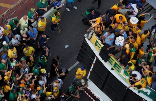 Multidão em verde e amarelo ao redor de um ônibus, com pessoas estendendo as mãos e olhando para cima.