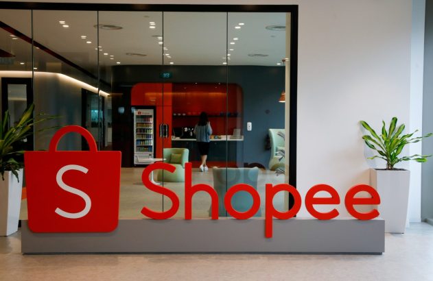 Entrada de escritório com o logo da Shopee. Através do vidro, uma mulher em um balcão e uma máquina de bebidas.
