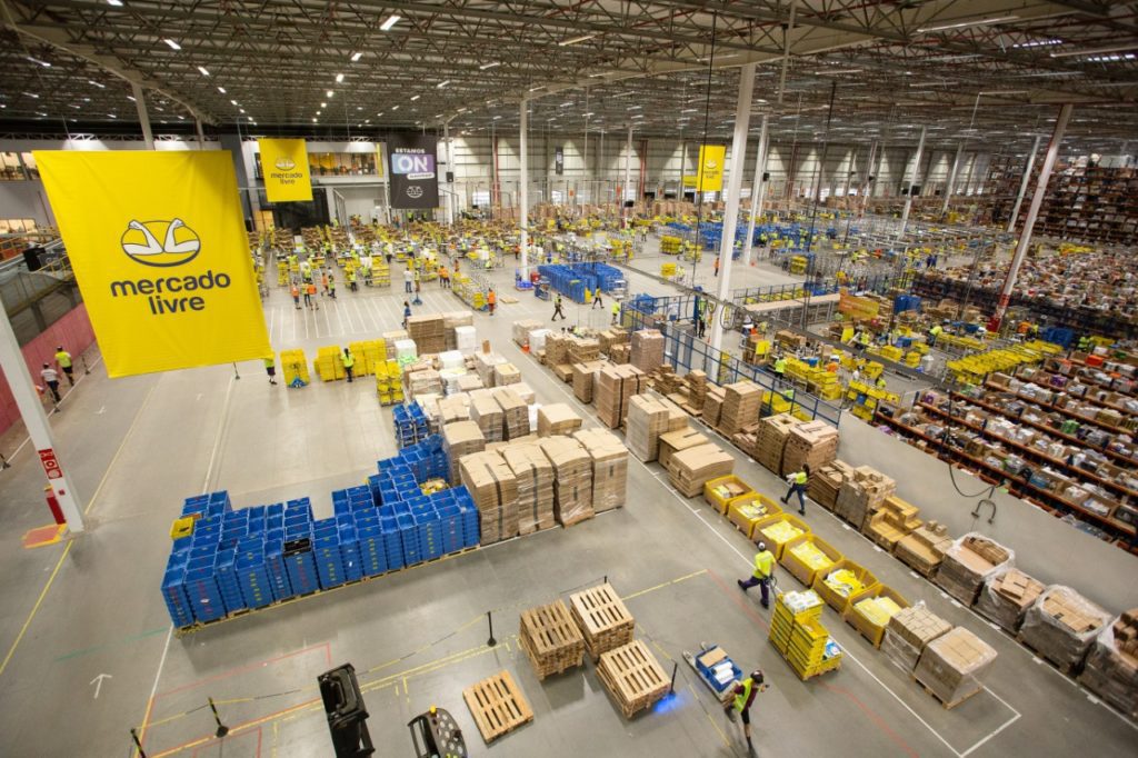 Grande centro de distribuição do Mercado Livre com trabalhadores movimentando caixas e produtos em diversas áreas de logística.