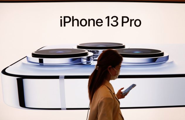 Mulher de máscara facial olhando o celular, passando em frente a um grande anúncio do iPhone 13 Pro.