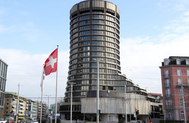 Um edifício cilíndrico imponente com janelas escuras, uma bandeira suíça e ciclistas em uma rua urbana.