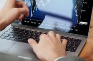 Pessoa usando laptop com tela exibindo gráfico financeiro. Uma mão aponta para os dados, a outra está no teclado.