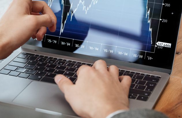 Pessoa usando laptop com tela exibindo gráfico financeiro. Uma mão aponta para os dados, a outra está no teclado.