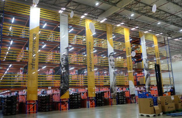 Centro de distribuição com prateleiras cheias de caixas, banners "infra.commerce" e uma empilhadeira em operação.