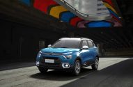 Citroën C3 azul com luzes acesas, em rua escura sob viaduto com mural colorido.