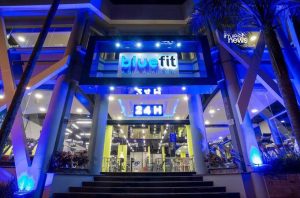 Entrada Bluefit à noite, iluminada em azul, com letreiros "A SUA ACADEMIA" e "24 H".