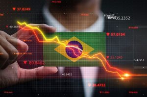 Mão sobre dados econômicos digitais da bandeira do Brasil, mostrando indicadores em queda. (88 caracteres)