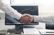 Homens se cumprimentam diante de monitor com gráfico de ações, fusões e aquisições decorativa