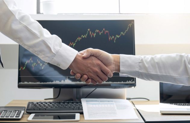 Homens se cumprimentam diante de monitor com gráfico de ações, fusões e aquisições decorativa