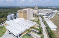 Vista aérea de complexo industrial com silos, fábrica, edifícios e palmeiras sob céu azul.