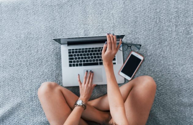 Pessoa sentada com pernas cruzadas usando laptop em superfície cinza. Óculos e celular ao lado.