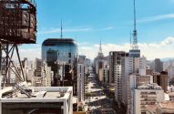 Vista aérea de avenida urbana ladeada por edifícios altos e torres, com carros na rua.